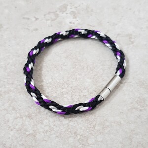 May include: A braided bracelet with black, white and purple cord, featuring a silver magnetic clasp.
