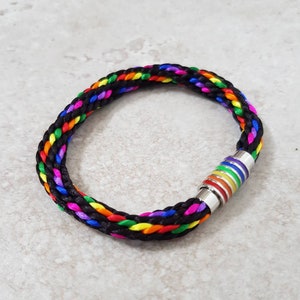 Puede incluir: Pulsera trenzada con un cordón negro y un arcoíris de colores tejidos. La pulsera tiene un cierre magnético plateado con rayas de arcoíris. La pulsera es un símbolo de orgullo e inclusión.