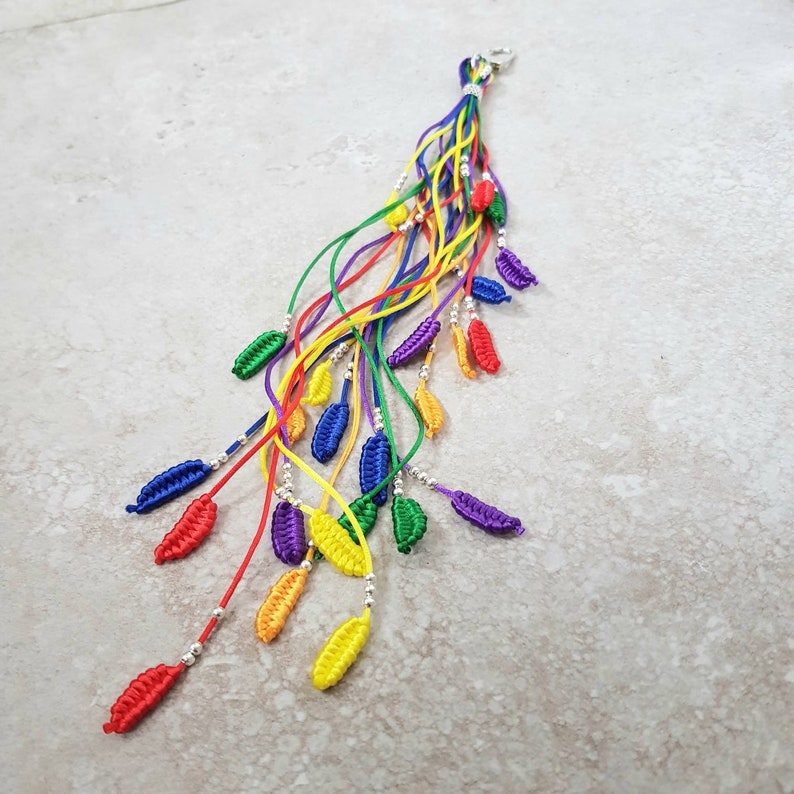 May include: A rainbow-colored string keychain with a silver charm. The keychain is made of multiple strands of string, each a different color of the rainbow. The strands are tied together at the top with a silver charm.