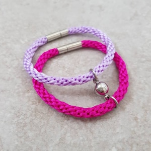 May include: Two braided bracelets, one pink and one lavender, with a silver magnetic clasp. The bracelets are made of a soft, woven material.