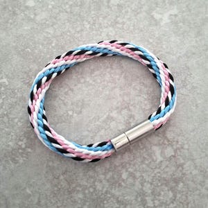以下が含まれることがあります： 黒、白、ピンク、水色の配色が特徴的な、編み込みブレスレット。シルバーカラーのマグネットクラスプが付いています。ブレスレットは円形で、柔らかい織り素材で作られているようです。