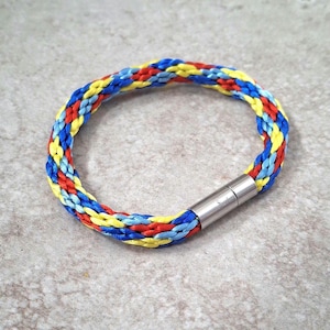 May include: A braided bracelet featuring a vibrant pattern of red, yellow, and blue threads. The bracelet is secured with a silver-colored magnetic clasp. The design is a colorful accessory.