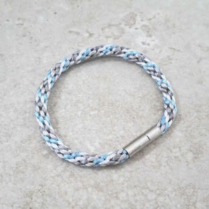 Puede incluir: Pulsera trenzada con hilos azules, blancos y grises. La pulsera tiene un cierre magnético plateado. Un accesorio sencillo y elegante. Circunferencia aproximada de 18 cm.