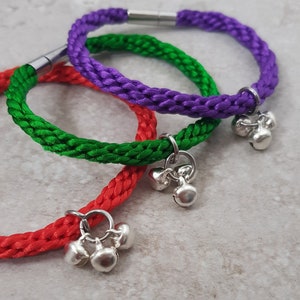 May include: Three braided cord bracelets with silver bell charms. The bracelets are red, green, and purple.
