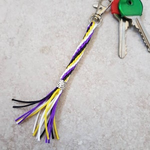 K&ouml;nnte beinhalten: Ein geflochtener Schl&uuml;sselanh&auml;nger in Lila, Wei&szlig; und Schwarz mit einer silbernen Perle und einem Quasten. Der Schl&uuml;sselanh&auml;nger ist an einem silbernen Schl&uuml;sselring mit zwei Schl&uuml;sseln befestigt.