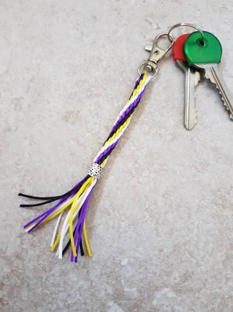 K&ouml;nnte beinhalten: Ein geflochtener Schl&uuml;sselanh&auml;nger in Lila, Wei&szlig; und Schwarz mit einer silbernen Perle und einem Quasten. Der Schl&uuml;sselanh&auml;nger ist an einem silbernen Schl&uuml;sselring mit zwei Schl&uuml;sseln befestigt.