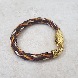 May include: Braided bracelet with a snake head clasp. The bracelet features a mix of brown, white, orange, and yellow threads. The snake head clasp is gold-colored and detailed.
