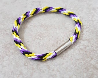 May include: A braided bracelet in the non-binary pride flag colours: purple, yellow, black, and white. It has a silver magnetic clasp. The design is a visible braid, showing the interwoven strands of colour.