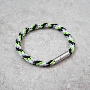 May include: Braided bracelet with a stainless steel magnetic clasp. The bracelet features a pattern of black, white, grey, and neon green cords. The bracelet is circular and rests on a grey surface.