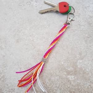May include: A colourful braided key chain with a silver bead and a tassel. The key chain is attached to two keys with red and green plastic covers.