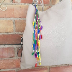 May include: A rainbow colored tassel keychain with silver beads and small blue, red, and yellow leaf charms attached to a light gray handbag.