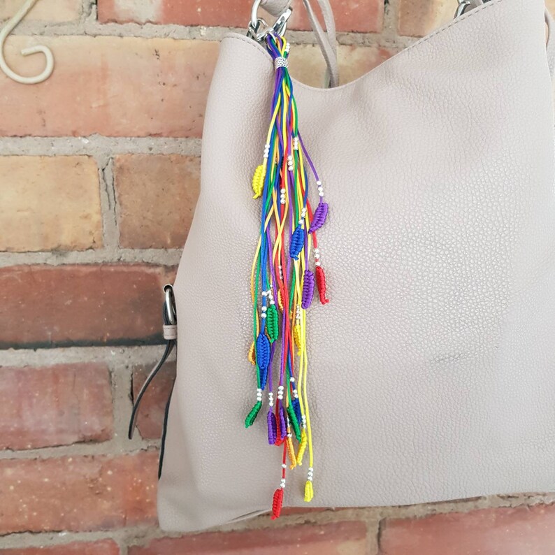 May include: A rainbow colored tassel keychain with silver beads and small blue, red, and yellow leaf charms attached to a light gray handbag.