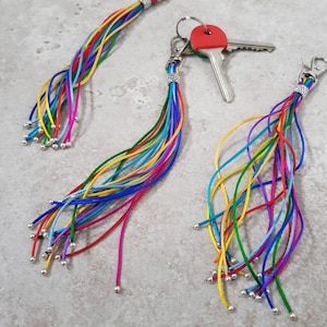 May include: Colorful keychains with multiple strands of vibrant cords in various colors, including red, yellow, blue, and green. Each keychain has a silver-colored clasp and small silver beads at the end of each cord. One keychain holds keys with a red key head.