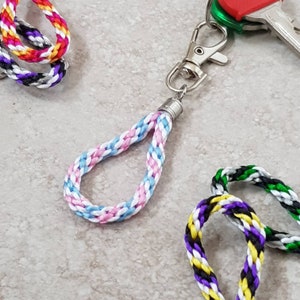 May include: A braided key chain with a light blue, pink and white colour scheme. The key chain has a silver clasp and is attached to a set of keys.