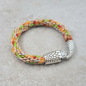 May include: A multi-coloured braided bracelet with a silver snake head clasp. The bracelet is made of beige, green, red, and orange threads.