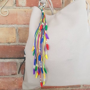 May include: A colorful rainbow tassel keychain with silver beads and small leaf-shaped charms. The keychain is attached to a light brown handbag.