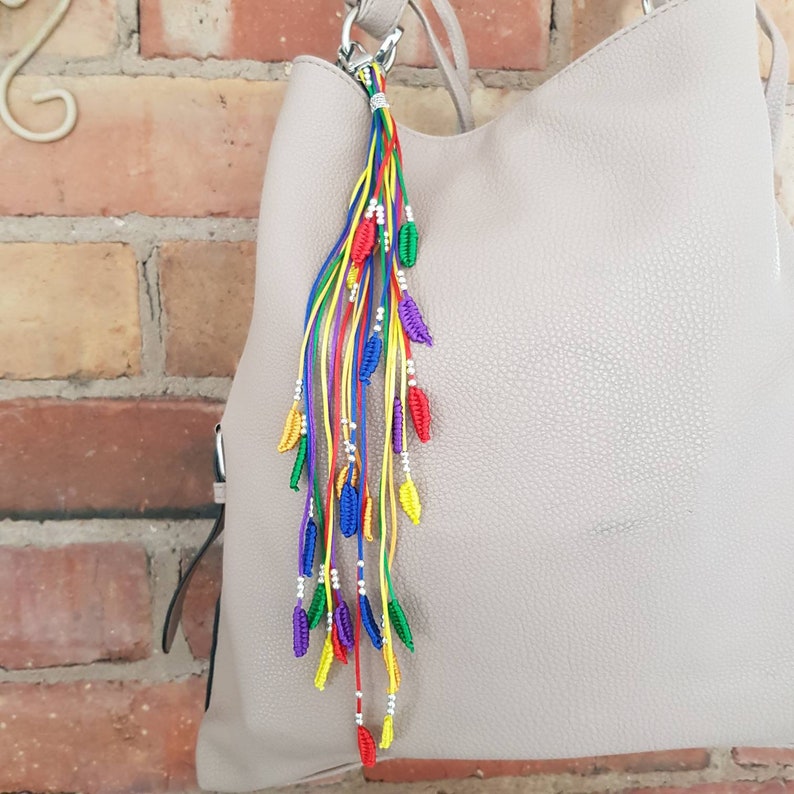 May include: A colorful rainbow tassel keychain with silver beads and small leaf-shaped charms. The keychain is attached to a light brown handbag.