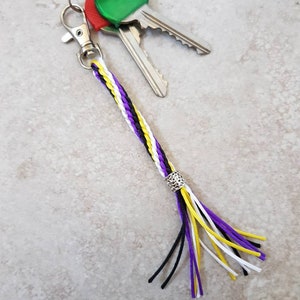 K&ouml;nnte beinhalten: Ein geflochtener Schl&uuml;sselanh&auml;nger in Lila, Wei&szlig; und Gelb mit einer silbernen Perle und einem schwarzen Quasten. Der Schl&uuml;sselanh&auml;nger ist an einem silbernen Schl&uuml;sselring mit zwei Schl&uuml;sseln befestigt.