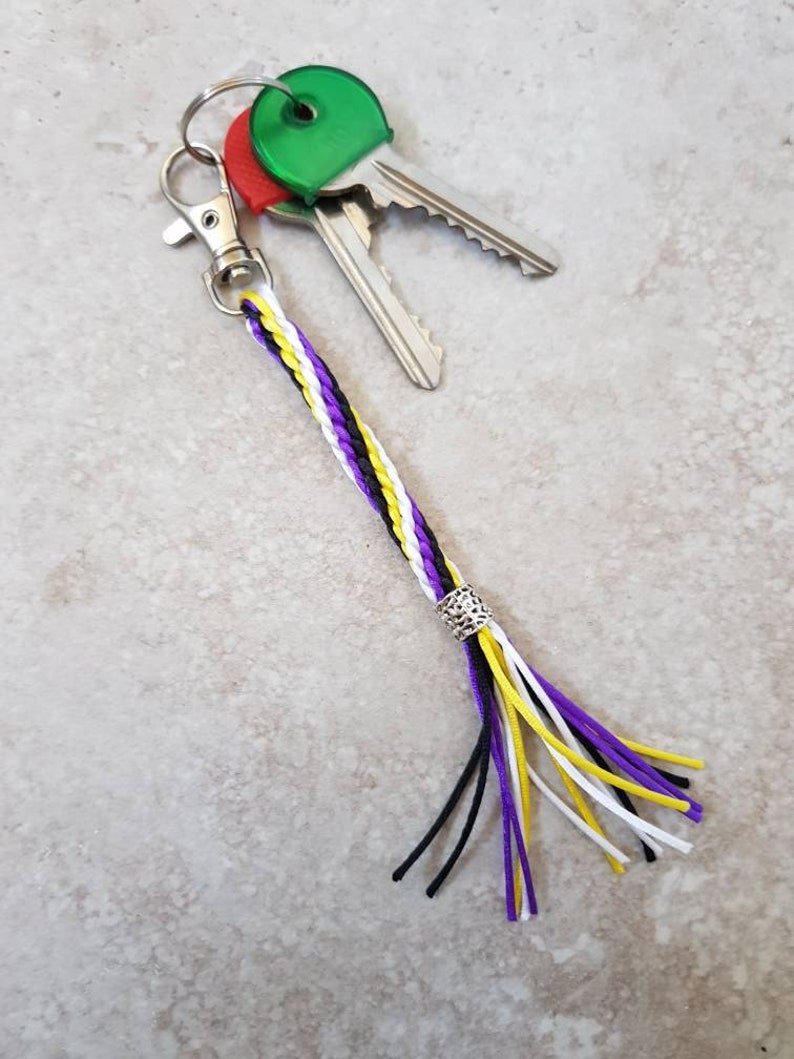 K&ouml;nnte beinhalten: Ein geflochtener Schl&uuml;sselanh&auml;nger in Lila, Wei&szlig; und Gelb mit einer silbernen Perle und einem schwarzen Quasten. Der Schl&uuml;sselanh&auml;nger ist an einem silbernen Schl&uuml;sselring mit zwei Schl&uuml;sseln befestigt.