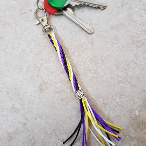 K&ouml;nnte beinhalten: Ein geflochtener Schl&uuml;sselanh&auml;nger mit einem lila, wei&szlig;en und gelben Quasten. Der Schl&uuml;sselanh&auml;nger hat einen silbernen Anh&auml;nger und ist an einem silbernen Schl&uuml;sselring mit drei Schl&uuml;sseln befestigt.