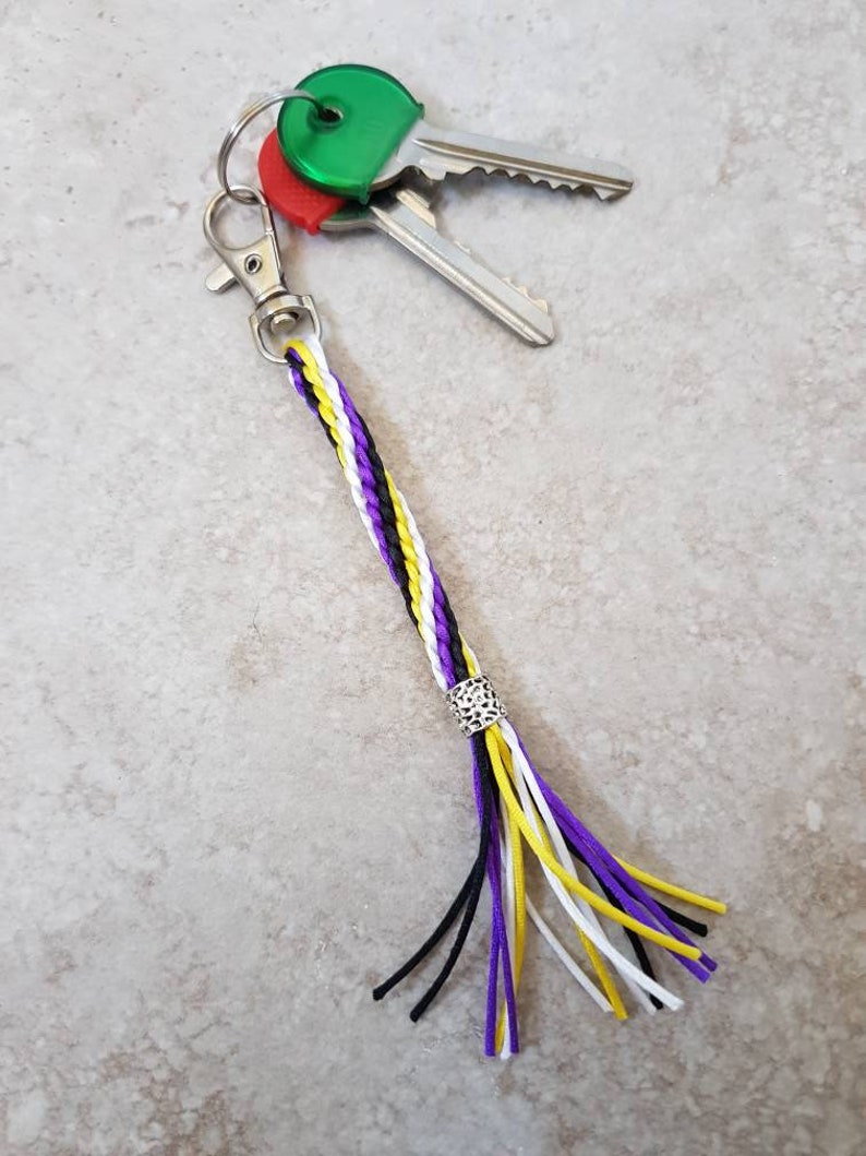 K&ouml;nnte beinhalten: Ein geflochtener Schl&uuml;sselanh&auml;nger mit einem lila, wei&szlig;en und gelben Quasten. Der Schl&uuml;sselanh&auml;nger hat einen silbernen Anh&auml;nger und ist an einem silbernen Schl&uuml;sselring mit drei Schl&uuml;sseln befestigt.