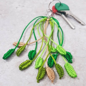 May include: A handmade macrame keychain featuring a cluster of green and tan leaf-shaped charms. The leaves vary in shades of green, with some tan accents. The keychain is attached to a silver clasp, with keys in the background.