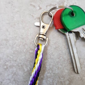 K&ouml;nnte beinhalten: Ein silberner Schl&uuml;sselanh&auml;nger mit einem geflochtenen Band in Schwarz, Wei&szlig;, Gelb und Lila. Der Schl&uuml;sselanh&auml;nger hat einen silbernen Clip und ist an zwei Schl&uuml;sseln mit roten und gr&uuml;nen Plastikkappen befestigt.