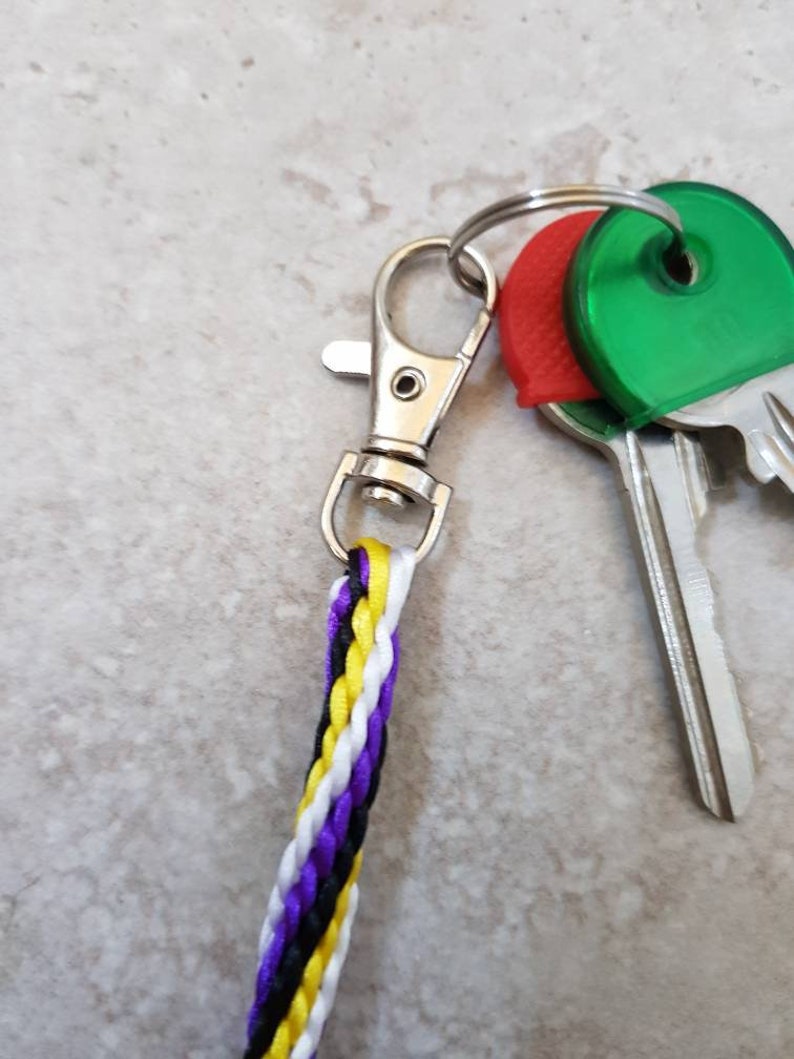 K&ouml;nnte beinhalten: Ein silberner Schl&uuml;sselanh&auml;nger mit einem geflochtenen Band in Schwarz, Wei&szlig;, Gelb und Lila. Der Schl&uuml;sselanh&auml;nger hat einen silbernen Clip und ist an zwei Schl&uuml;sseln mit roten und gr&uuml;nen Plastikkappen befestigt.