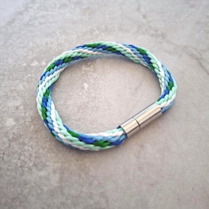 May include: Braided bracelet in shades of blue, green, and white, with a silver-tone magnetic clasp. The bracelet is circular in shape. A stylish accessory for the wrist.