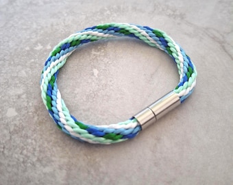 May include: Braided bracelet featuring a mix of blue, green, and white cords. The bracelet has a silver-colored magnetic clasp. The design is a circular shape, suitable for wrist wear.