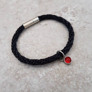 May include: Black braided bracelet with a silver clasp and a small, round, red charm. The bracelet is made of a thick, woven material. The charm is a bright red colour. The bracelet is a simple, elegant design.