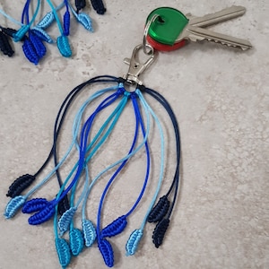 May include: A keychain with a silver clasp and multiple blue macrame tassels. The tassels vary in shades of blue, from light to dark, and are attached to a silver clasp holding two keys.
