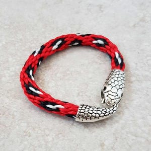 Puede incluir: Una pulsera trenzada con un patrón rojo, negro y blanco. La pulsera presenta un cierre de cabeza de serpiente plateada. Un accesorio único.