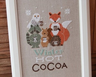 May include: A framed cross-stitch design featuring a fox, owl, raccoon, squirrel, and hedgehog, all holding mugs of hot cocoa. The text "Winter Hot Cocoa" is stitched below the animals.