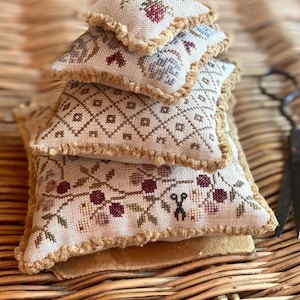 Può includere: Una pila di quattro piccoli cuscini quadrati ricamati con uno sfondo beige e motivi floreali marroni, rossi e blu. I cuscini hanno una frangia marrone attorno ai bordi.