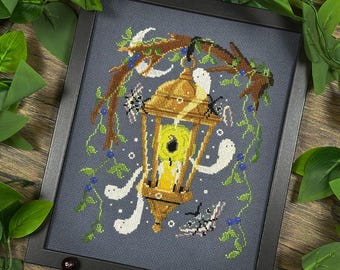May include: Cross-stitch art in a black frame. The design features a golden lantern with a lit candle, surrounded by white ghost figures, spiders, and a moth. The background is a dark gray with green vines and blue berries.