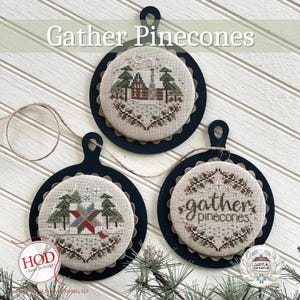 May include: Three round, black framed ornaments with a white background and a cross-stitch design. The top ornament features a winter scene with a cabin and trees. The middle ornament features a red and green quilt block. The bottom ornament features a wreath with the text "gather pinecones".