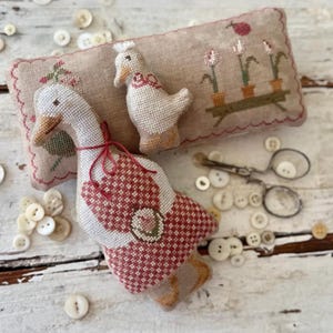 May include: Cross-stitched goose and duck ornaments with red and white patterned wings, a small embroidered pillow, and vintage scissors. The scene is set on a white wooden surface, with scattered buttons. Measurements are in cm.