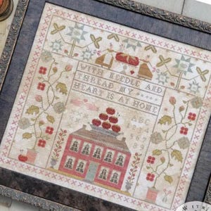 Può includere: Campione a punto croce incorniciato con la scritta "WITH NEEDLE AND THREAD MY HEART IS AT HOME". Il disegno presenta una casa rossa, elementi floreali e bordi decorativi. La cornice è marrone scuro.