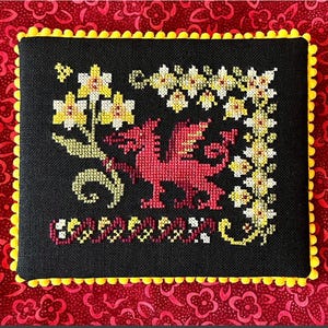 Pode incluir: Arte de ponto cruz em tecido preto com um dragão vermelho, flores amarelas e brancas e uma borda decorativa. É emoldurado com uma borda de pompom amarelo e colocado sobre um fundo floral vermelho.