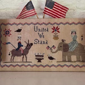 Op de afbeelding: Een kruissteek-sampler met de tekst "United We Stand" in het midden. De sampler toont een ezel, een olifant, een vogel, een bloem en twee Amerikaanse vlaggen.