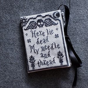 Op de afbeelding: Een wit naaldvilt met zwarte borduurwerk dat "Here lie dead My needle and thread" zegt. Het naaldvilt is vastgebonden met een zwart lint.