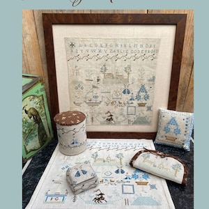 May include: A cross-stitch sampler featuring the text "Lvlie Godefroid" and an alphabet chart. The sampler is framed in a dark wood frame and is surrounded by other cross-stitch projects, including a small cushion, a box, and a cylindrical container.