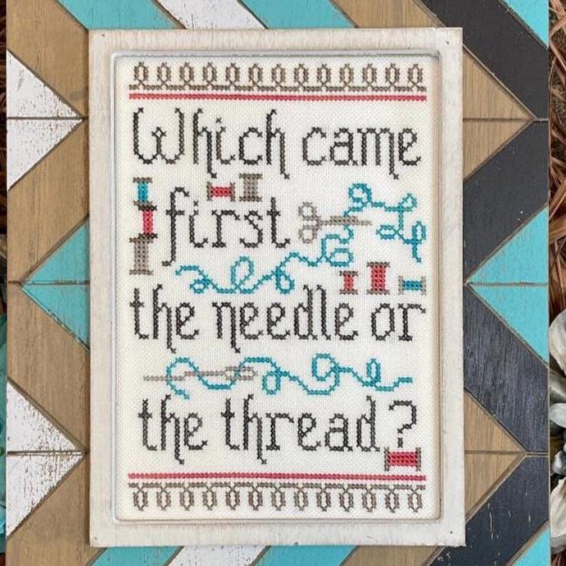 May include: Cross-stitch artwork in a white frame, featuring the question "Which came first, the needle or the thread?" The design includes thread spools, a needle, and teal thread. The frame is set against a colourful geometric background.