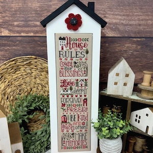 Peut inclure: Un cadre en bois blanc en forme de maison contenant une broderie au point de croix avec le texte House Rules, Count Your Blessings, Love Deeply, Forgive Freely, Be Kind, Laugh Often et Make Memories, surmonté d'une fleur rouge au crochet.
