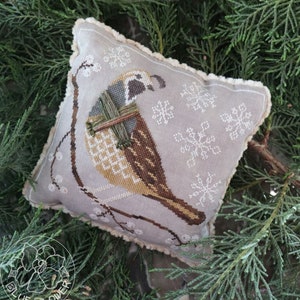 May include: A gray and white embroidered pillow with a brown and white quail perched on a branch. The quail is holding a bundle of twigs in its beak. The background is a light gray with white snowflakes.
