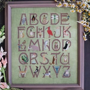 May include: Framed cross-stitch sampler showing the alphabet, each letter with an animal illustration. The letters and animals are stitched in brown thread on a pale green background. The frame is light brown.