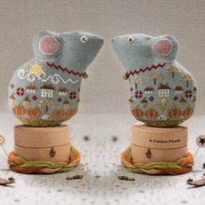 May include: Two gray fabric mouse figurines with embroidered details, including houses and trees, perched on small wooden spools. Each mouse has pink ears and a decorative collar. The spools are set on a woven base.
