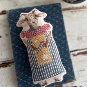 May include: A handmade goat-shaped fabric doll with a detailed cross-stitch design. The doll features a tan face, a red and white patterned top, and a striped blue and white dress. It holds a yellow bag with a star and a red and white design.