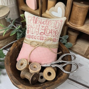 May include: A small, pink and beige decorative pillow embroidered with the text "Abide in My Love" in pink. The pillow is tied with twine and sits in a wooden bowl with thread spools and vintage scissors. Wooden shelves and decorative hearts are in the background.